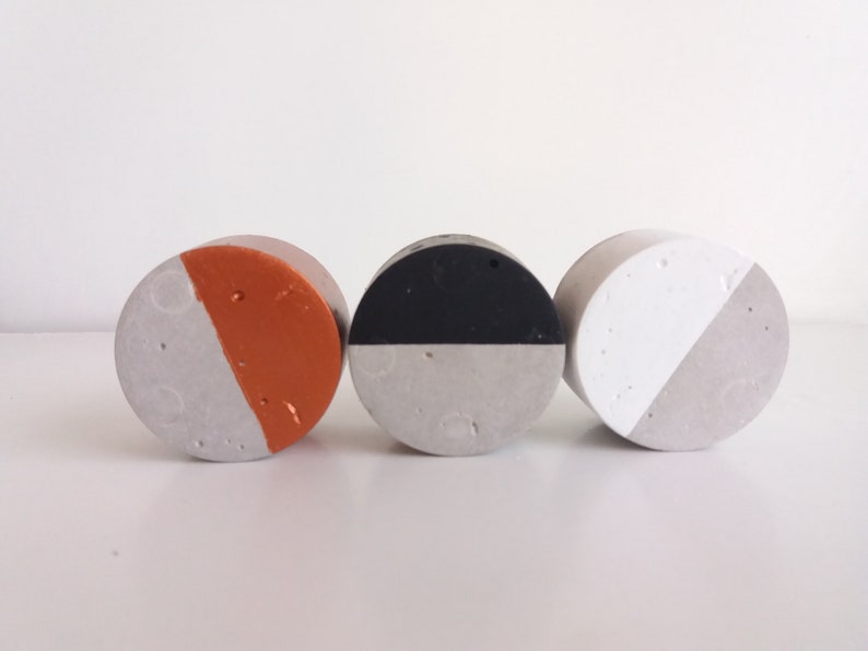 May include: Three round concrete coasters with a half-circle design. The coasters are painted in different colours: orange, black, and white.
