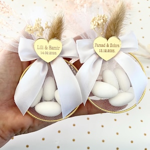 May include: Two gold-rimmed favour containers filled with white sweets, decorated with white bows, tulle, and dried flowers. Each has a gold heart with names and a date: "Lili & Samir 14.02.2026" and "Farzad & Zohra 13.12.2025".