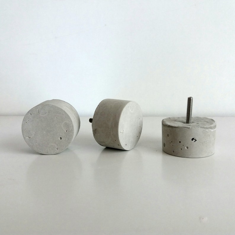 May include: Three concrete furniture legs, two round and one with a screw top. The legs are a light grey colour and have a rough, textured surface.