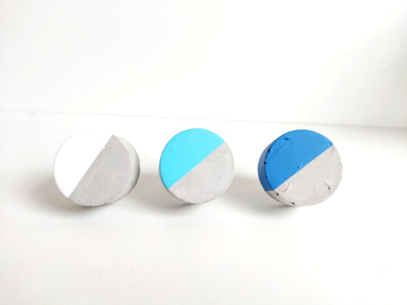 May include: Three round concrete knobs with a half-circle of paint on each. The knobs are painted white, light blue, and dark blue.