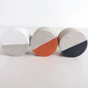 May include: Three round concrete coasters with a white top and a different coloured bottom. The coasters are grey, orange, and black.