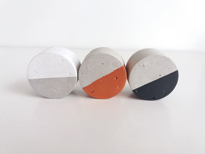 May include: Three round concrete coasters with a white top and a different coloured bottom. The coasters are grey, orange, and black.