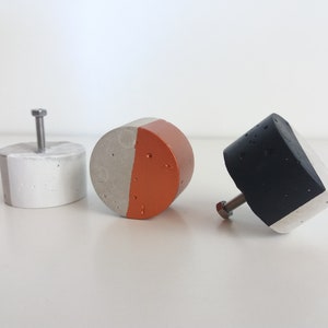 May include: Three concrete cabinet knobs with a half-round design. The knobs are painted in different colours: white and grey, grey and orange, and black and white.