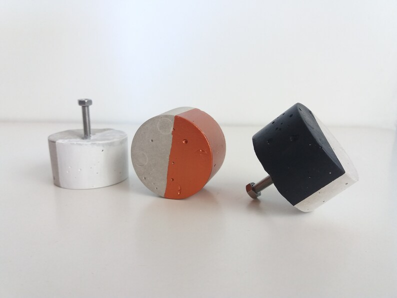 May include: Three concrete cabinet knobs with a half-round design. The knobs are painted in different colours: white and grey, grey and orange, and black and white.