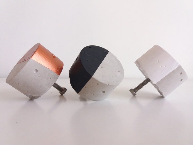 May include: Three concrete cabinet knobs with different finishes. One is painted black and white, one is painted copper and white, and one is all white.