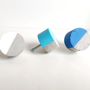 May include: Three concrete knobs with a half-circle of blue paint on each. One knob is attached to a metal screw. The knobs are on a white surface.