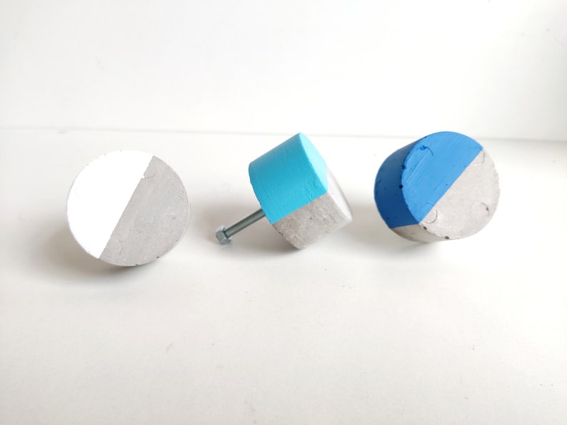 May include: Three concrete knobs with a half-circle of blue paint on each. One knob is attached to a metal screw. The knobs are on a white surface.