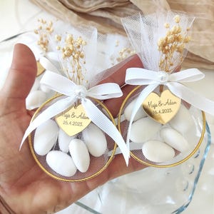 Op de afbeelding: Twee huwelijksbedankzakjes. Elke zak heeft een goudomrande cirkel gevuld met witte snoepjes, een witte tule top, een wit lint en een goudkleurig hartvormig label met tekst. Gedroogde bloemen en een parel accentueren de bovenkant.