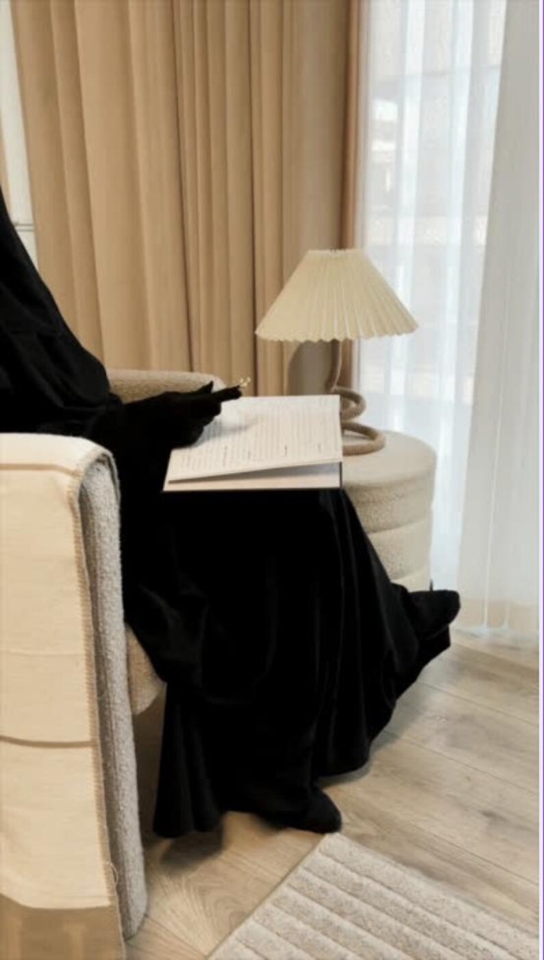 Op de afbeelding: Een persoon in een zwart kledingstuk zit in een beige fauteuil, met een open boek op schoot. Een cr&egrave;mekleurige lamp met een geplooide kap staat op een kleine, ronde poef. De kamer heeft neutrale gordijnen en een lichte houten vloer.