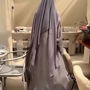 Op de afbeelding: Een persoon draagt een lange, vloeiende, lichtpaarse abaya en bijpassende hoofdbekleding. De abaya heeft geborduurde details. De persoon staat in een goed verlichte kamer met tafels en stoelen, mogelijk een restaurant.