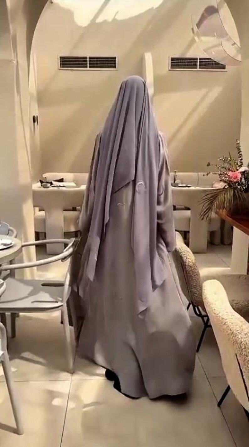 Op de afbeelding: Een persoon draagt een lange, vloeiende, lichtpaarse abaya en bijpassende hoofdbekleding. De abaya heeft geborduurde details. De persoon staat in een goed verlichte kamer met tafels en stoelen, mogelijk een restaurant.