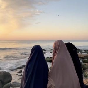 Op de afbeelding: Drie figuren in vloeiende kleding staan op een strand en kijken bij zonsondergang naar de oceaan. De figuren zijn gehuld in marineblauwe, mauve en zwarte stoffen. De lucht is een verloop van oranje en blauw, met zachtjes op de kust rollende golven.