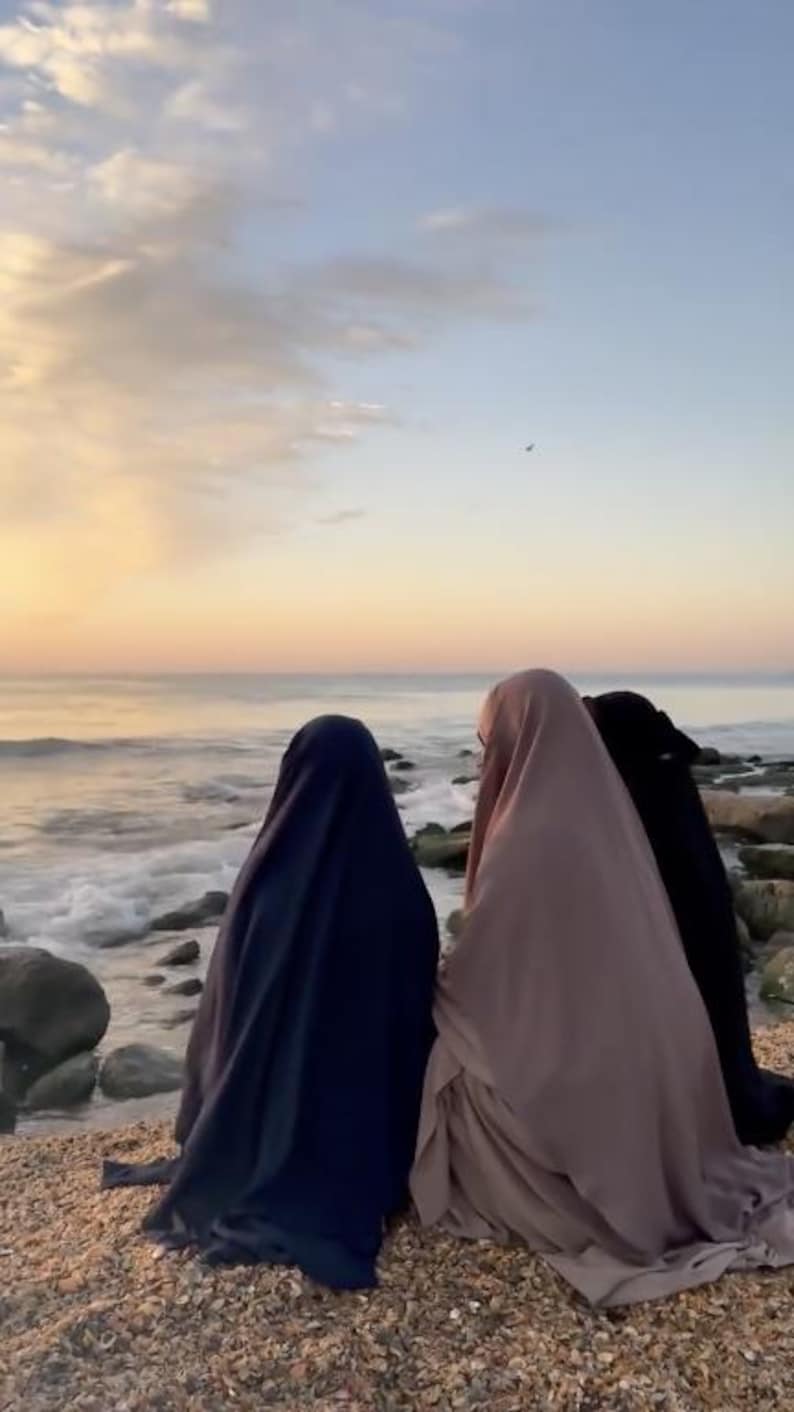 Op de afbeelding: Drie figuren in vloeiende kleding staan op een strand en kijken bij zonsondergang naar de oceaan. De figuren zijn gehuld in marineblauwe, mauve en zwarte stoffen. De lucht is een verloop van oranje en blauw, met zachtjes op de kust rollende golven.