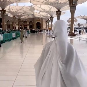 Op de afbeelding: Een persoon in witte kleding loopt door een grote, open structuur met talrijke witte, paraplu-achtige luifels. De grond is geplaveid met witte tegels en andere mensen zijn in de verte zichtbaar.