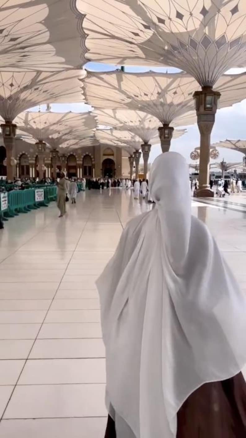 Op de afbeelding: Een persoon in witte kleding loopt door een grote, open structuur met talrijke witte, paraplu-achtige luifels. De grond is geplaveid met witte tegels en andere mensen zijn in de verte zichtbaar.