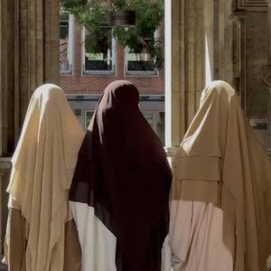 Op de afbeelding: Drie personen gekleed in gedrapeerde kleding in tinten beige, bruin en wit staan onder een gewelfde stenen structuur. De sc&egrave;ne wordt overspoeld door zonlicht, met een glimp van een gebouw en bomen op de achtergrond.