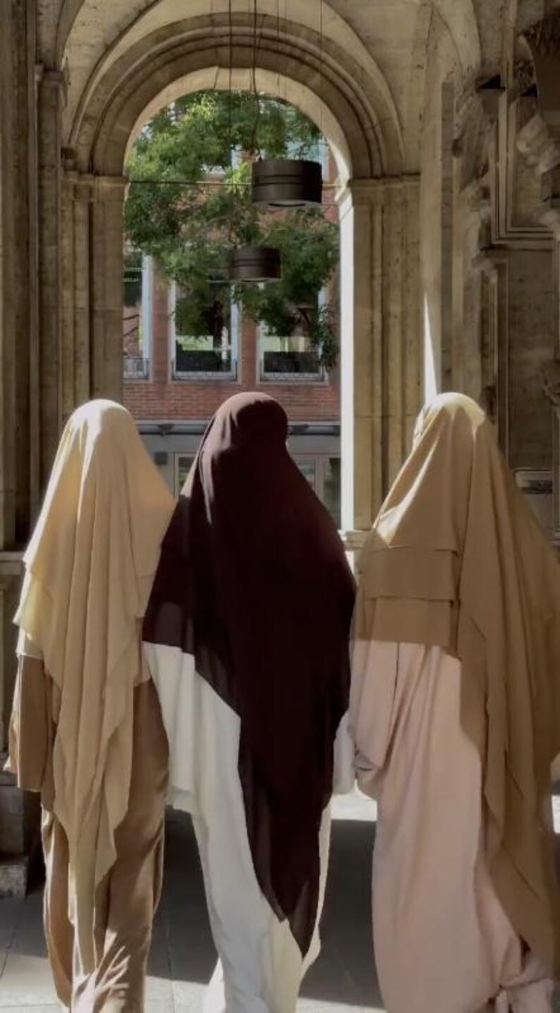 Op de afbeelding: Drie personen gekleed in gedrapeerde kleding in tinten beige, bruin en wit staan onder een gewelfde stenen structuur. De sc&egrave;ne wordt overspoeld door zonlicht, met een glimp van een gebouw en bomen op de achtergrond.