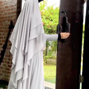 Op de afbeelding: Een persoon die een lichtgrijze hijab en jurk draagt, opent een donkerbruine houten deur. De hijab heeft gelaagde details en de mouwen hebben een zwarte bies. De achtergrond omvat een bakstenen muur en groen.