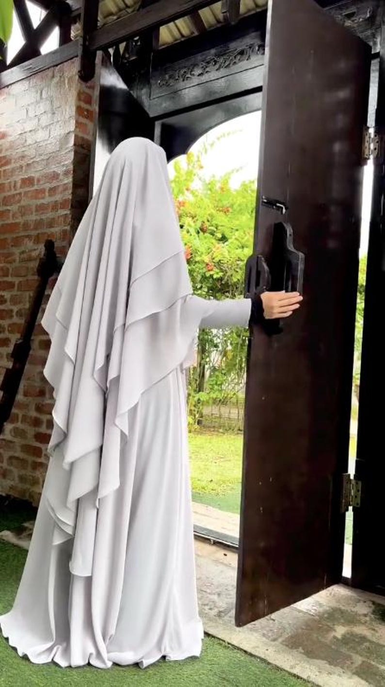 Op de afbeelding: Een persoon die een lichtgrijze hijab en jurk draagt, opent een donkerbruine houten deur. De hijab heeft gelaagde details en de mouwen hebben een zwarte bies. De achtergrond omvat een bakstenen muur en groen.