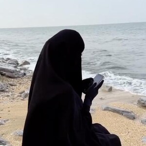 Op de afbeelding: Een persoon in een zwarte niqab zit op een rots bij de zee. De persoon houdt een smartphone vast, met de zee en de lucht op de achtergrond. De niqab bedekt het hoofd en lichaam.