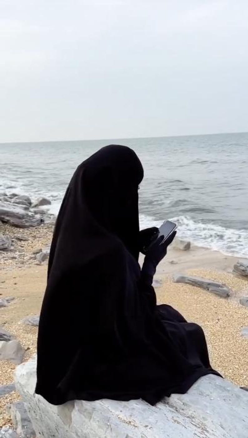 Op de afbeelding: Een persoon in een zwarte niqab zit op een rots bij de zee. De persoon houdt een smartphone vast, met de zee en de lucht op de achtergrond. De niqab bedekt het hoofd en lichaam.