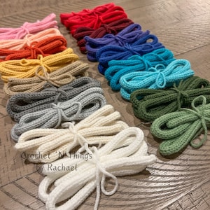 May include: A collection of crocheted cords in various colors, including pink, red, maroon, blue, teal, green, gray, and white. The cords are arranged in a row on a wooden surface. The text "Crochet 'N Things by Rachael" is visible in the lower left corner of the image.