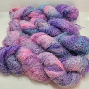 May include: A skein of hand-dyed yarn in shades of pink, purple, and blue. The yarn has a soft, fuzzy texture and appears to be a blend of fibers. The colors blend together, creating a gradient effect. Ideal for knitting or crochet projects.