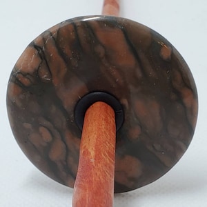 May include: A spinning wheel whorl with a brown and orange marbled design. The whorl is attached to a wooden spindle.