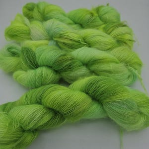 May include: A collection of three skeins of fuzzy, lightweight yarn in vibrant shades of lime green, chartreuse, and pale yellow. The yarn features a soft, haloed texture with subtle color variations throughout each twisted, hand-wound bundle.