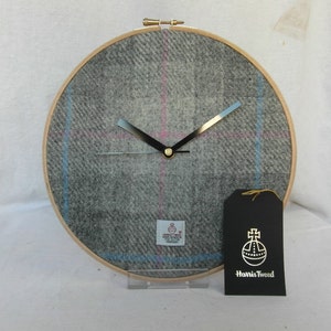 May include: A gray and blue plaid clock with black hands and a gold center. The clock is in a wooden hoop and has a black tag with the text "Harris Tweed" and a gold logo.