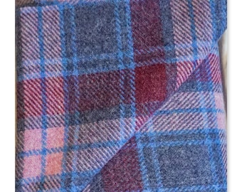 May include: Close-up of a folded plaid fabric in shades of blue, burgundy, and pink. The fabric has a classic tartan pattern with intersecting lines creating a grid-like design. The texture appears soft and possibly woolen.