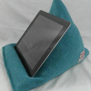 May include: A teal blue tweed tablet stand with a tablet resting on top. The stand is shaped like a wedge and has a label that says "Handmade in Scotland".
