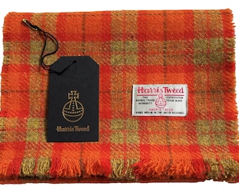 May include: Orange and green plaid Harris Tweed scarf with fringed edges. The Harris Tweed label is attached to the scarf. The label reads "Harris Tweed", "The Harris Tweed Authority", "Certification Trade Mark", and "Hand Woven in the Outer Hebrides".