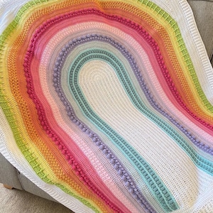 May include: A rainbow crochet baby blanket with a white background and a crocheted border. The blanket features a rainbow arch design with different coloured stripes and textured stitches.