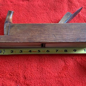 May include: A vintage wooden hand plane with a steel blade. The plane is brown and measures approximately 10 inches long. The tool is designed for woodworking and is placed on a red surface.