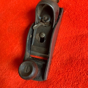 May include: A vintage metal block plane with a dark finish. The plane has a curved top, a blade adjustment lever, and a wooden knob. The tool is designed for woodworking and is set against a red background.