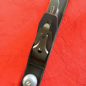 May include: A gray hand plane with a light blue knob and metal blade. The plane is set against a red background. The tool is designed for woodworking and shaping wood surfaces.