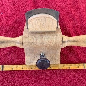 May include: A wooden drawknife with a metal blade and two wooden handles. The blade is set in a wooden frame with the text "CME 06". A wooden ruler is placed below the drawknife. The background is red.