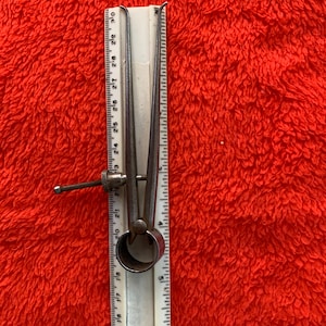 Puede incluir: Un calibre de metal plateado con una regla blanca marcada en pulgadas. El calibre está abierto y la regla está marcada de 3 a 30 pulgadas.