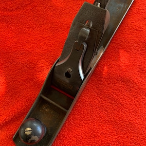 May include: A vintage hand plane, featuring a dark metal body with a wooden knob and handle. The blade is visible, and the tool rests on a red surface. This woodworking tool is designed for shaping and smoothing wood surfaces.