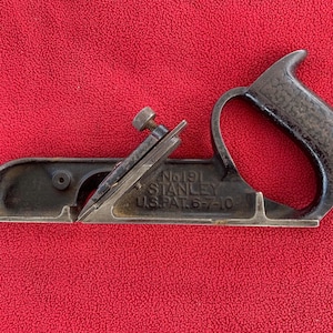 May include: A vintage Stanley No. 191 block plane, featuring a dark handle and body with a blade secured by a screw. The tool has the text "No. 191 STANLEY U.S.A. PAT. 6-7-10" etched on its side. The plane is set against a red background.