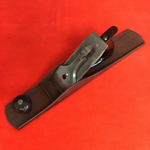 May include: A black wooden hand plane with a metal blade and a knob on the front. The plane is on a red surface.