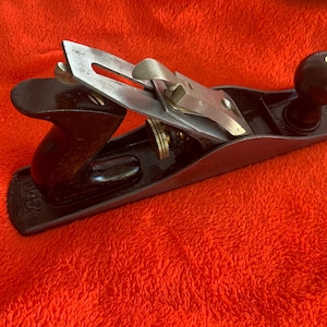 May include: A vintage hand plane with a black body, a dark wooden handle, and a silver blade. The plane rests on a bright red surface, showcasing its classic design and craftsmanship. The tool is designed for woodworking.