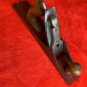 May include: A vintage hand plane with a dark brown wooden body and a metal blade. The plane features a wooden handle and knob, and the metal blade is secured with a lever cap. The tool is designed for woodworking and smoothing surfaces.