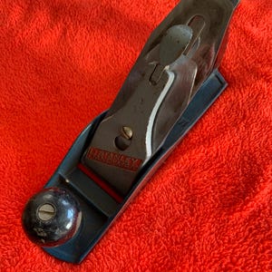 May include: A vintage Stanley Handyman smoothing plane. The plane has a blue body, a black knob, and a silver blade. The text "STANLEY HANDYMAN" is visible on the blade. The plane is set against a bright red background.