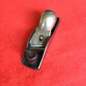 May include: A metal block plane featuring a silver-toned body with a rounded palm handle and a textured grip area. The tool includes a dark brown circular knob at the front and a central screw mechanism for adjusting the blade depth and alignment.
