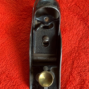 Op de afbeelding: Een vintage handvlak, met een donkere metalen behuizing, een getextureerde zoolplaat en een messing afstelknop. Deze houtbewerkingsgereedschap heeft een klassieke, versleten uitstraling. Het vlak ligt op een felrode achtergrond. Ongeveer 15 cm lang.