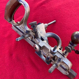 May include: A vintage metal woodworking plane with a wooden handle and a blade. The plane is silver and brown, with adjustable knobs and screws. The words "MADE IN U.S.A." are visible on the side. The tool is designed for shaping and smoothing wood surfaces.