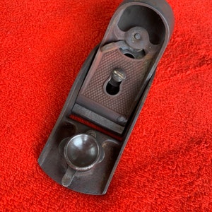May include: A vintage metal hand plane with a dark brown finish. The plane has a metal knob and a lever on the top.