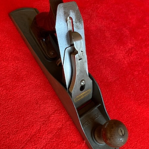 May include: A vintage Craftsman hand plane, featuring a dark gray metal body with a wooden handle and knob. The blade is silver, and the tool is marked "MADE IN USA". The plane rests on a red surface.